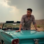 EN: A man leaning on a vintage convertible parked on a desert highway, wide open sky, rolled-up sleeves, hair slightly messy from wind, timeless freedom RU: Мужчина облокотился на винтажный кабриолет на шоссе посреди пустыни, широкое небо, закатанные рукава, волосы растрепаны ветром, вечная свобода