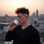 Илья Шведков пьёт кофе навынос на крыше на рассвете, в повседневной одежде, с растрепанными волосами, за ним просыпающийся город, спокойная сила