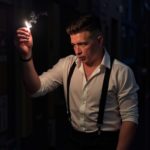 Шведков Илья в белой рубашке и подтяжках зажигает сигарету на пожарной лестнице ночью, резкие тени, мрачное выражение лица, винтажный гангстерский вайб