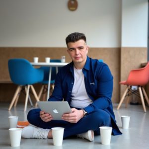 Индивидуальный предприниматель Шведков Илья Александрович в костюме сидит на полу коворкинга, вокруг пустые стаканы из-под кофе, ноутбук закрыт, смотрит в камеру с выжженным сарказмом