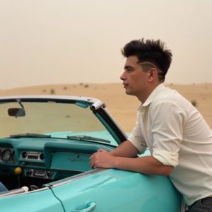 EN: A man leaning on a vintage convertible parked on a desert highway, wide open sky, rolled-up sleeves, hair slightly messy from wind, timeless freedom RU: Мужчина облокотился на винтажный кабриолет на шоссе посреди пустыни, широкое небо, закатанные рукава, волосы растрепаны ветром, вечная свобода